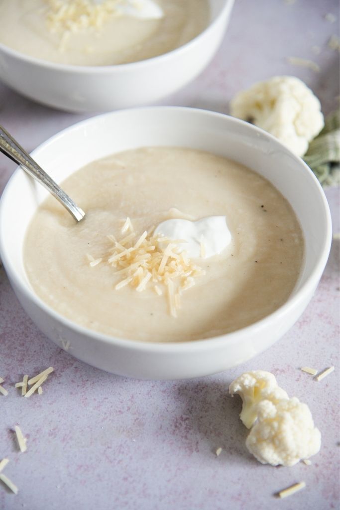 a bowl of soup with a spoon it, there is a piece of cauliflower in front of the bowl 
