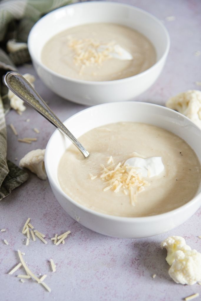 a bowl of soup with a spoon in it, you can see another bowl of soup and some pieces of cauliflower behind the first bowl 