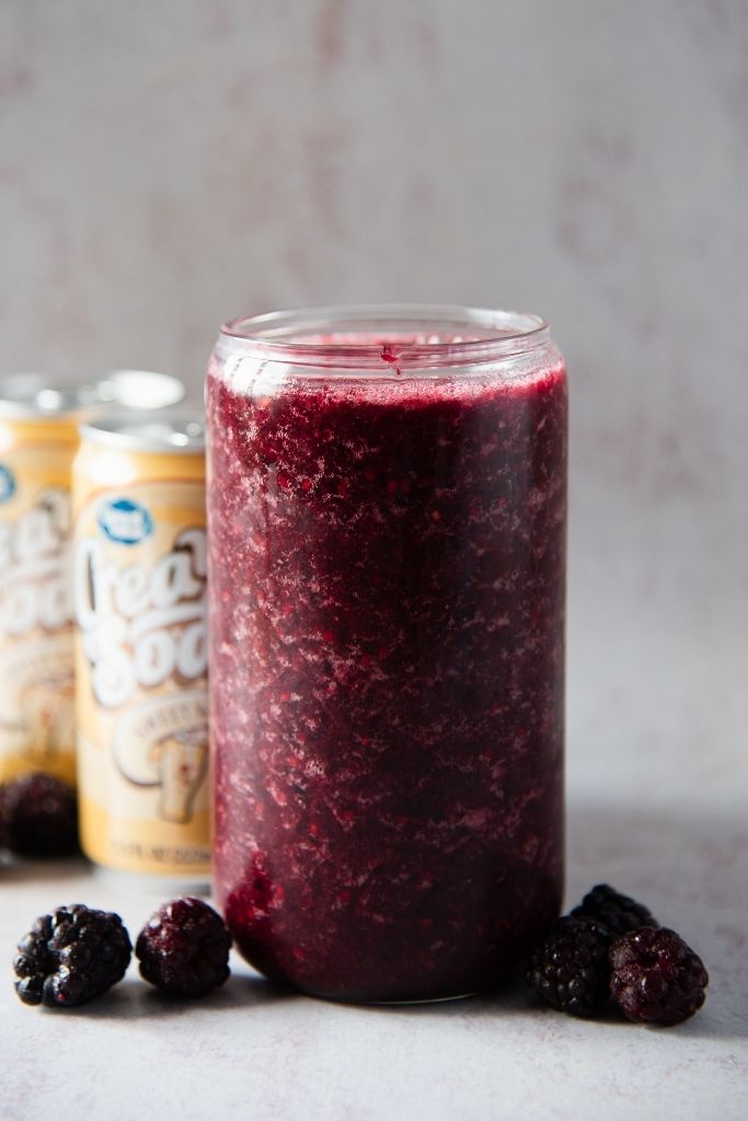 a close up of a blackberry slushy, there are two cans of soda behind the slushy 