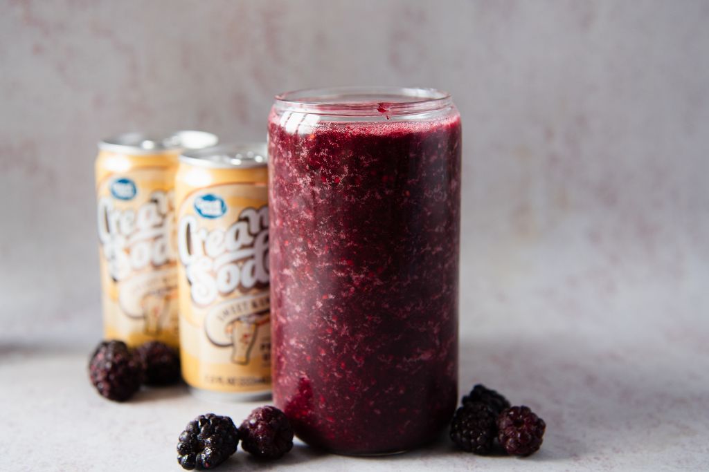 a blackberry slushy with blackberries around it, there is also two cans of soda next to it 
