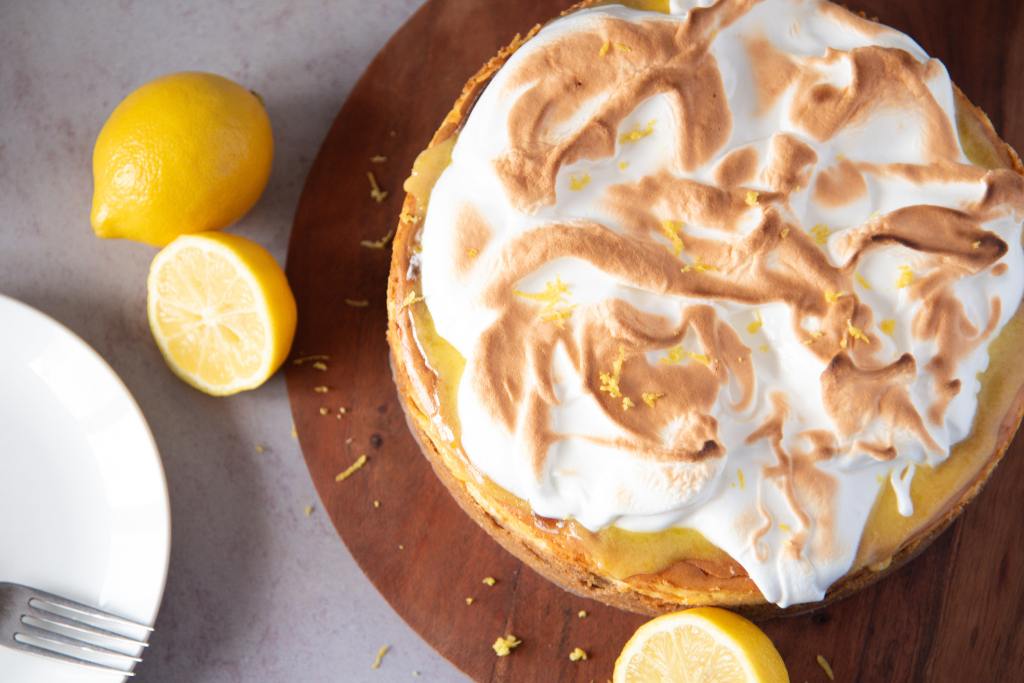an uncut cheesecake with lemons next to it, there is also a plate with a fork on it, next to the cheesecake