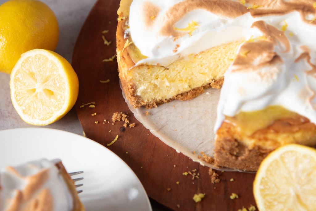 a cheesecake with a slice taken out of it, there are some lemons around the cheesecake and you can see part of piece of cheesecake on a plate off to the side