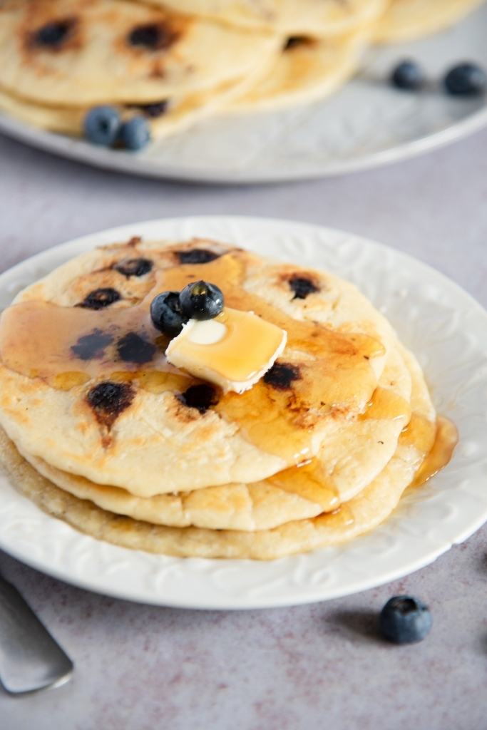 a close-up of a plate of blueberry pancakes, there is butter and syrup on top of the pancakes