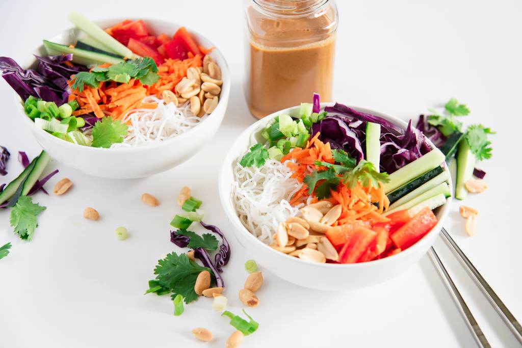 two bowls of spring roll salad, there is a jar of peanut dressing next to them, and some chopped veggies on the table around them