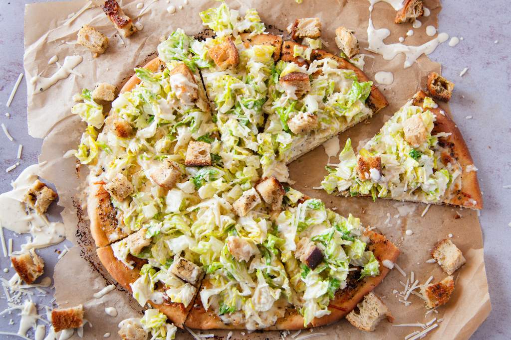 a cut Caesar salad pizza with, one slice has been pulled away from the rest of the pizza