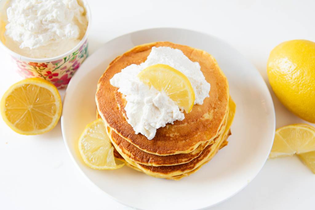 a plate of lemon pancakes with whipped cream on top, there is also a dish of whipped cream, some lemons and lemon slices around the plate 