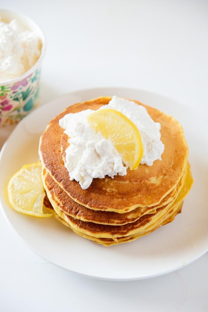 a stalk of pancakes with whipped cream and lemon slices on top. You can also see a dish of whipped cream next to the pancakes 