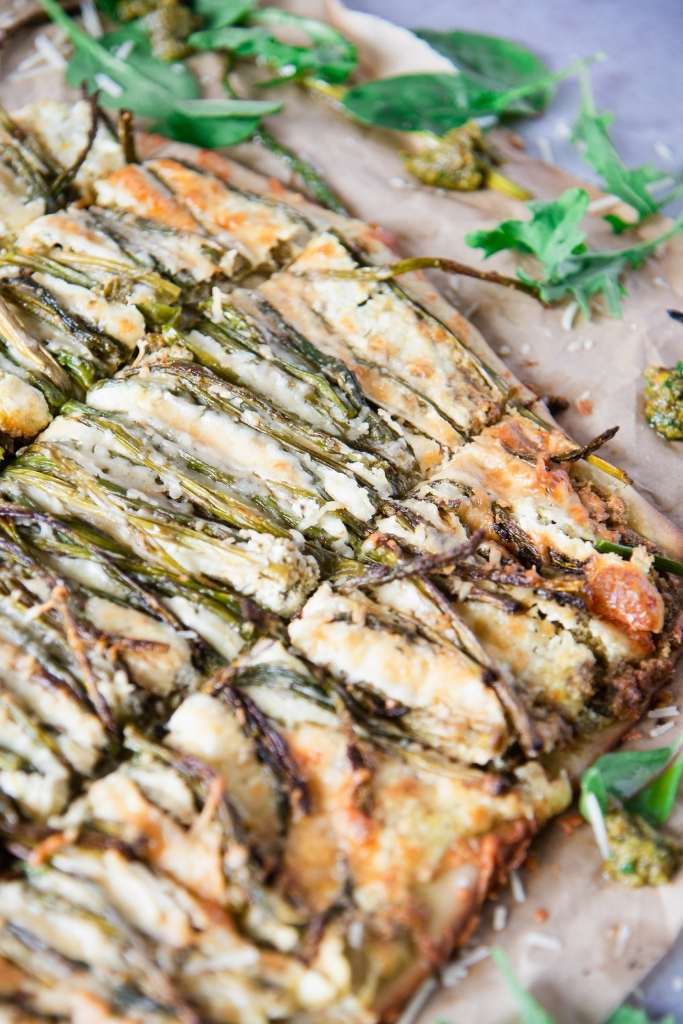 a close-up of a cut pizza with asparagus on it, there is some pesto and greens by the pizza too.