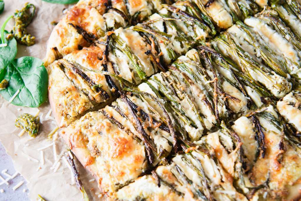 a cut pizza with asparagus on it, there is pesto and shredded cheese spilled around the pizza 