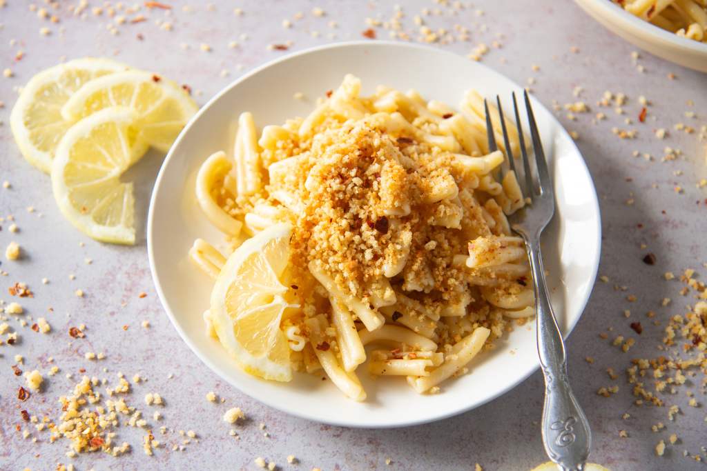 a plate of pasta, there is also a fork and lemon slices on the plate, there are also breadcrumbs and lemon slices around the plate