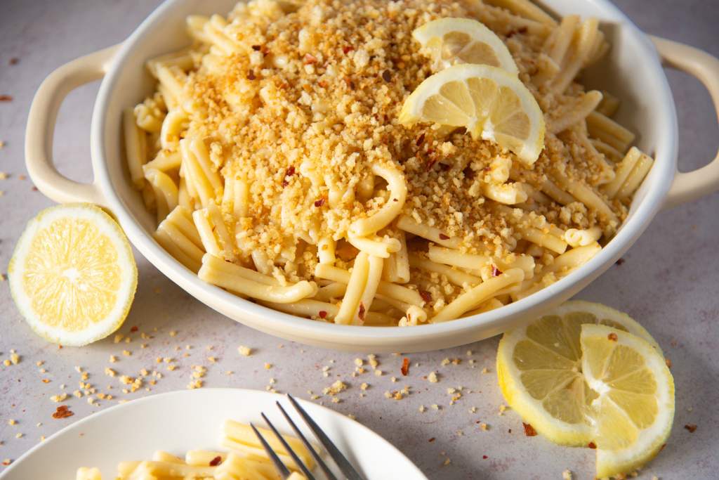 a dish of pasta with breadcrumbs on top, there are lemon slices next to the dish, and you can see a plate of pasta as well