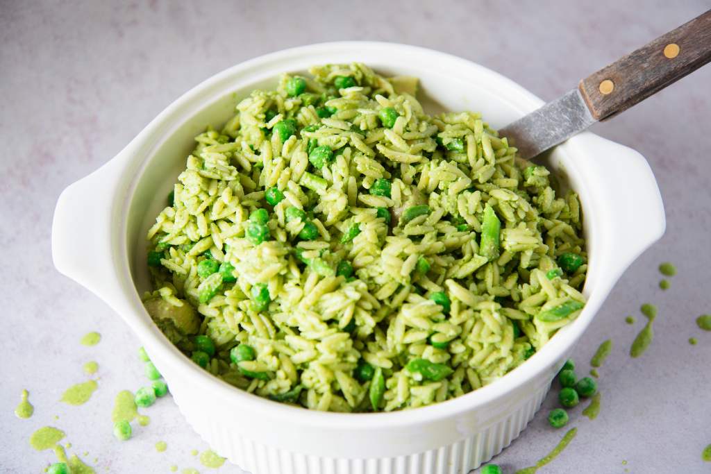 a bowl of pasta salad with a spoon in it, there is some green dressing and some peas spilled around the bowl