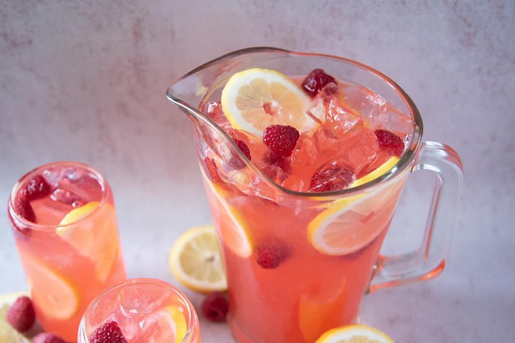 a pitcher of lemonade with two glasses of lemonade next to it, there are also some raspberries and lemon slices set around them