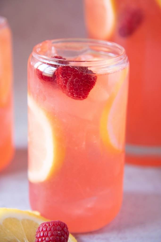 a close up of a glass of lemonade, there is a lemon slice and a fresh raspberry next to the glass