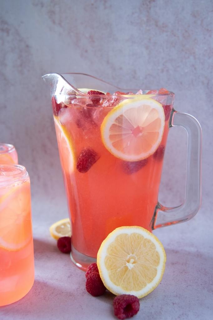 a pitcher of lemonade, there is a half a lemon and some fresh raspberries next to it 