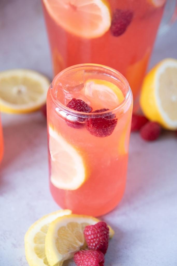 a glass of lemonade with lemon slices and fresh raspberries floating in it. You can also see a pitcher of lemonade behind the glass