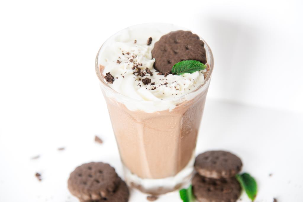a shake with whipped cream and a cookie on top, there is also cookies and mint leaves next to the shake 
