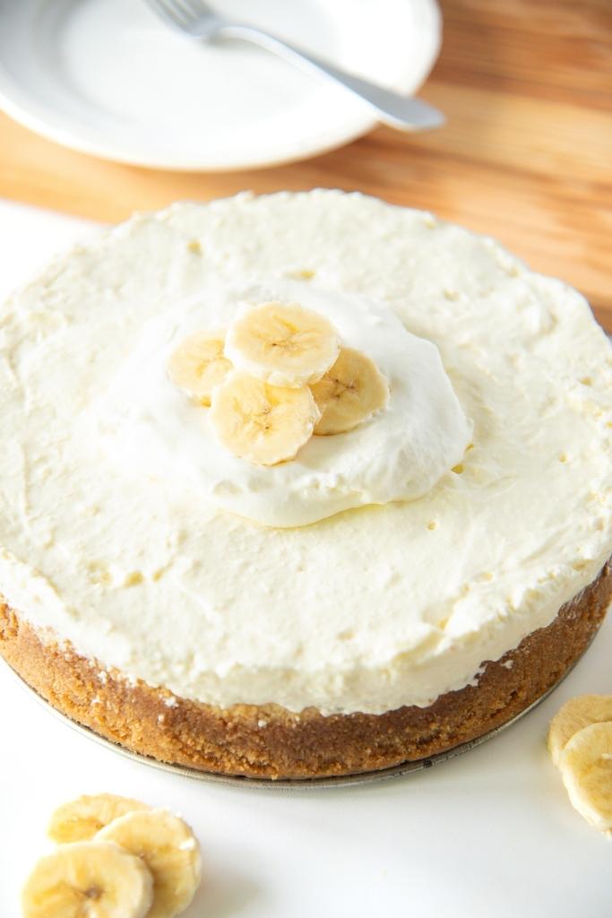 a banana pudding cheesecake with whipped cream and sliced bananas on it, you can also see a plate and fork behind the cheesecake.