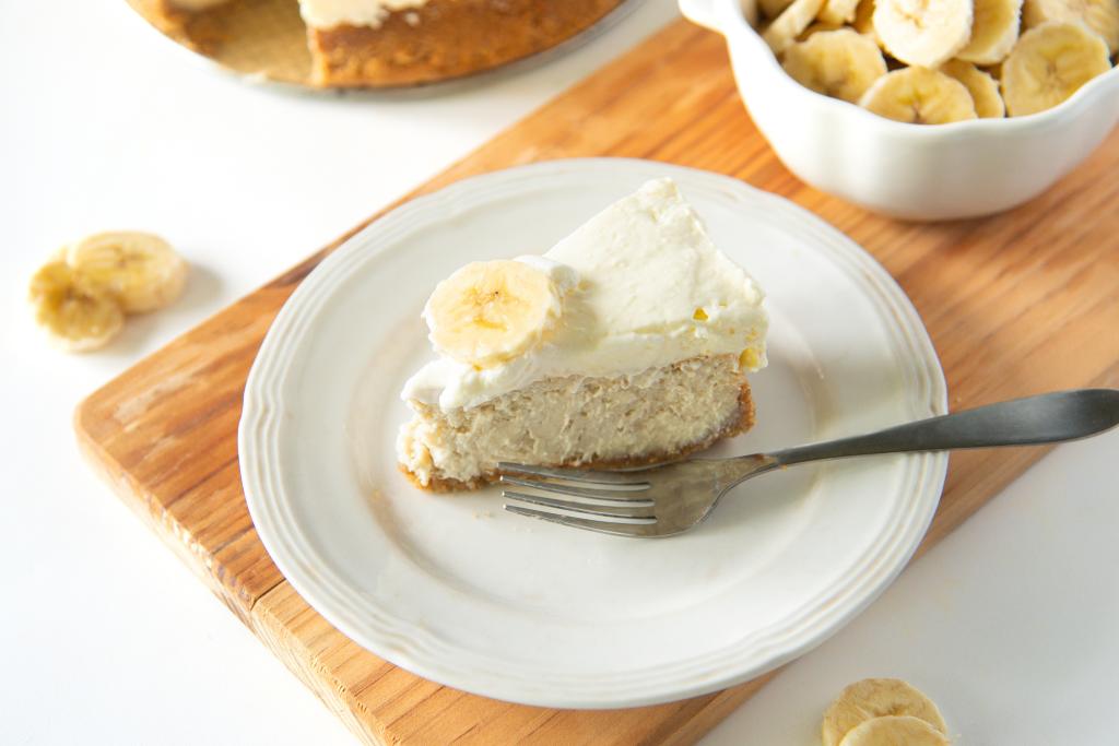 a slice of cheesecake and a fork on a plate, you can also see a dish of banana slices behind the plate 