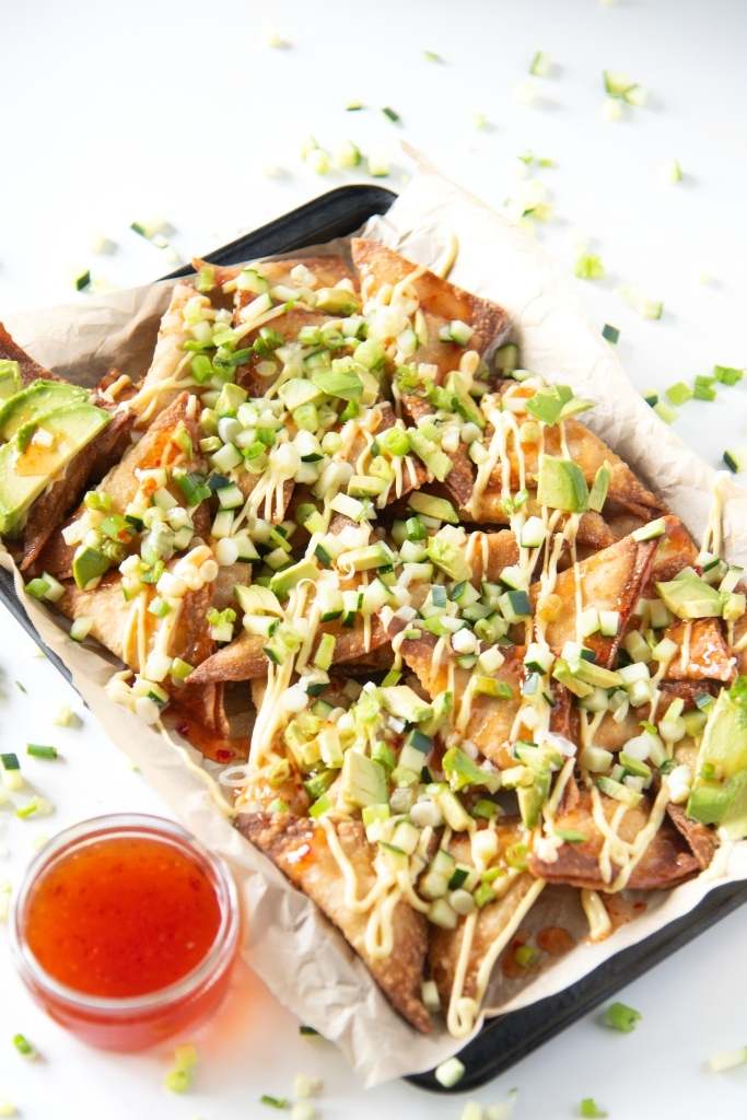 a tray of rangoon nachos with a dish of sweet chili sauce next to it. There is also some cucumber pieces and green onions around the tray 