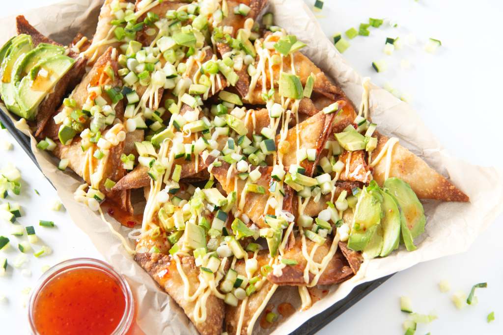 a tray of rangoon nachos with sliced avocado on them, there is also a dish of sweet chili sauce next to the tray