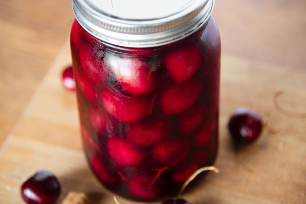 a jar of pickled cherries with fresh cherries and a cinnamon stick next to it 