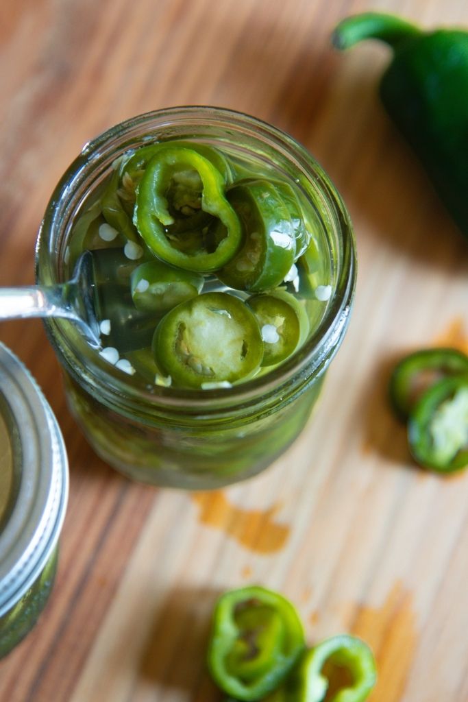 a jar of jalapenos with a fork in it, there also a whole jalapeno and some slices next to the jar