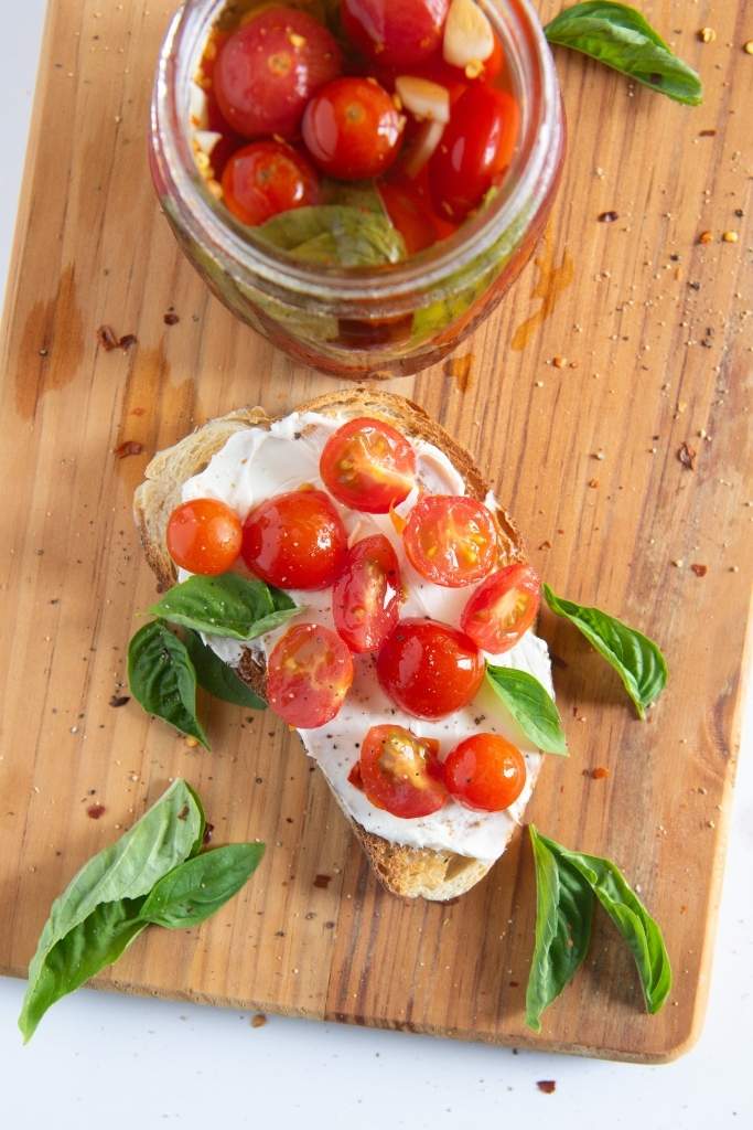 a piece of toast with pickled tomatoes on it, there is also a jar of tomatoes and a fresh basil next to the toast