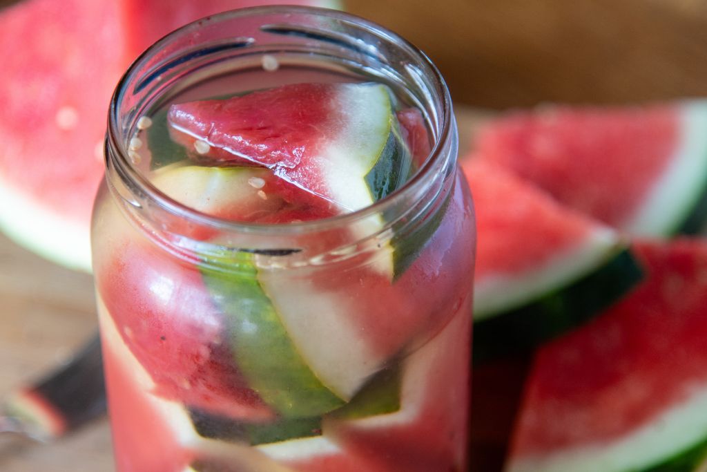 an open jar of pickled watermelon, there are some pieces of fresh watermelon behind the jar