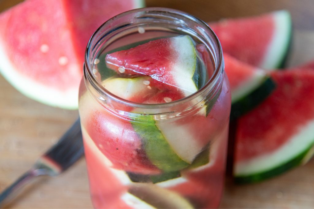 a jar of pickled watermelon there is fresh watermelon pieces and a fork next to the jar