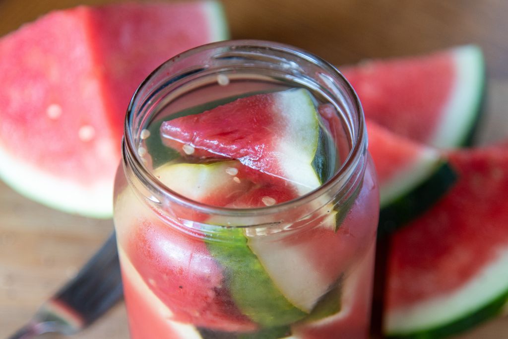 a jar of pickled watermelon, there are watermelon pieces and a fork next to the jar