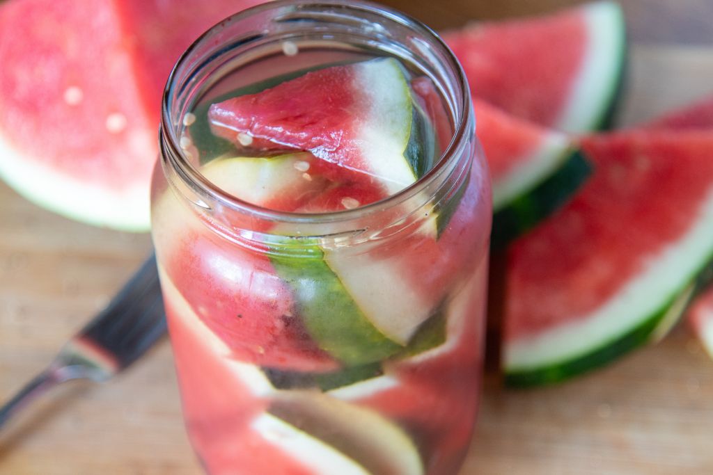 a jar of pickled watermelon, there is a fork and some fresh watermelon next to the jar 