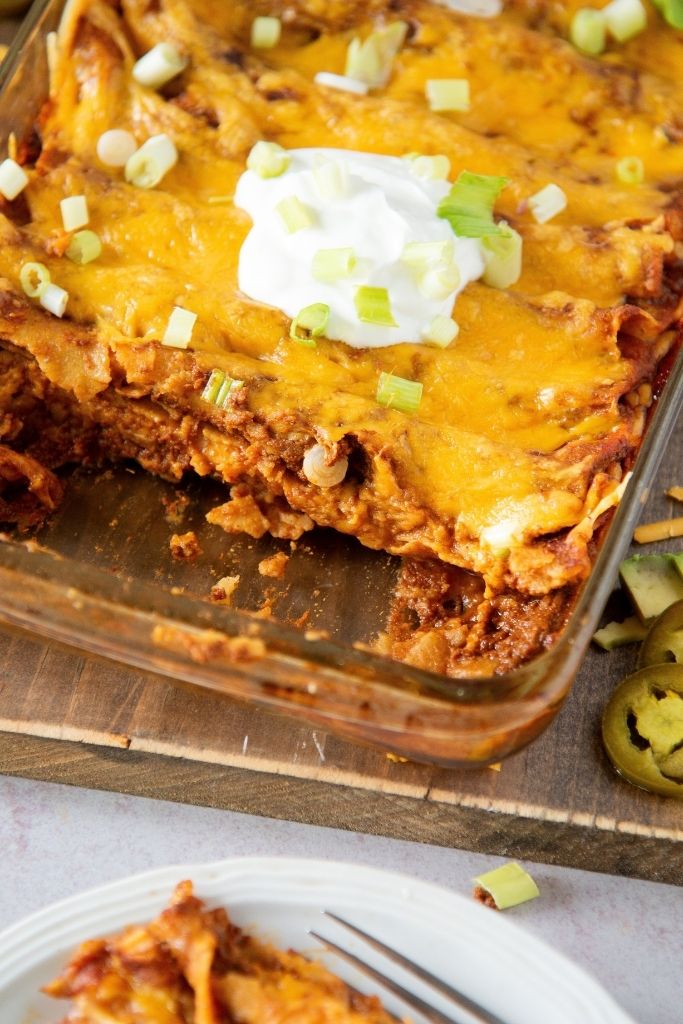 a pan of enchiladas with some taken out of it, there is also sour cream and green onions on top of the enchiladas