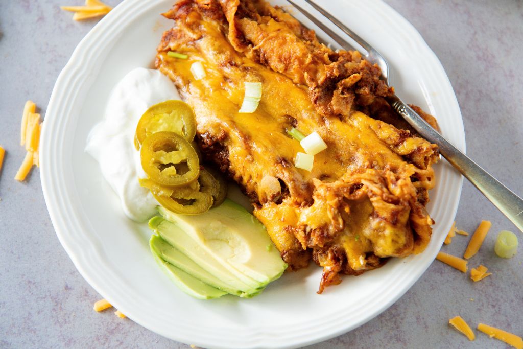 a plate of enchiladas with a fork on it, there also sour cream, sliced avocado and pickled jalapenos on the side